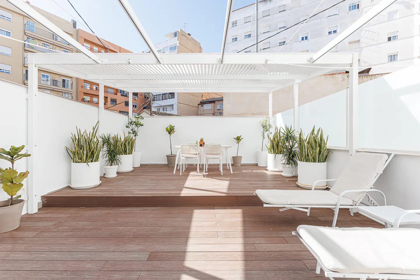 A rooftop patio with a white pergola, wooden deck, white furniture, and potted plants.