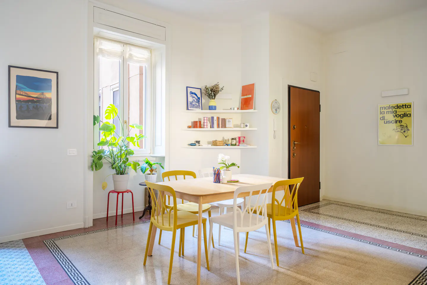 Bright dining room with a light wood table surrounded by yellow and white chairs. Shelves with books and decor are on the wall. A framed print hangs nearby.