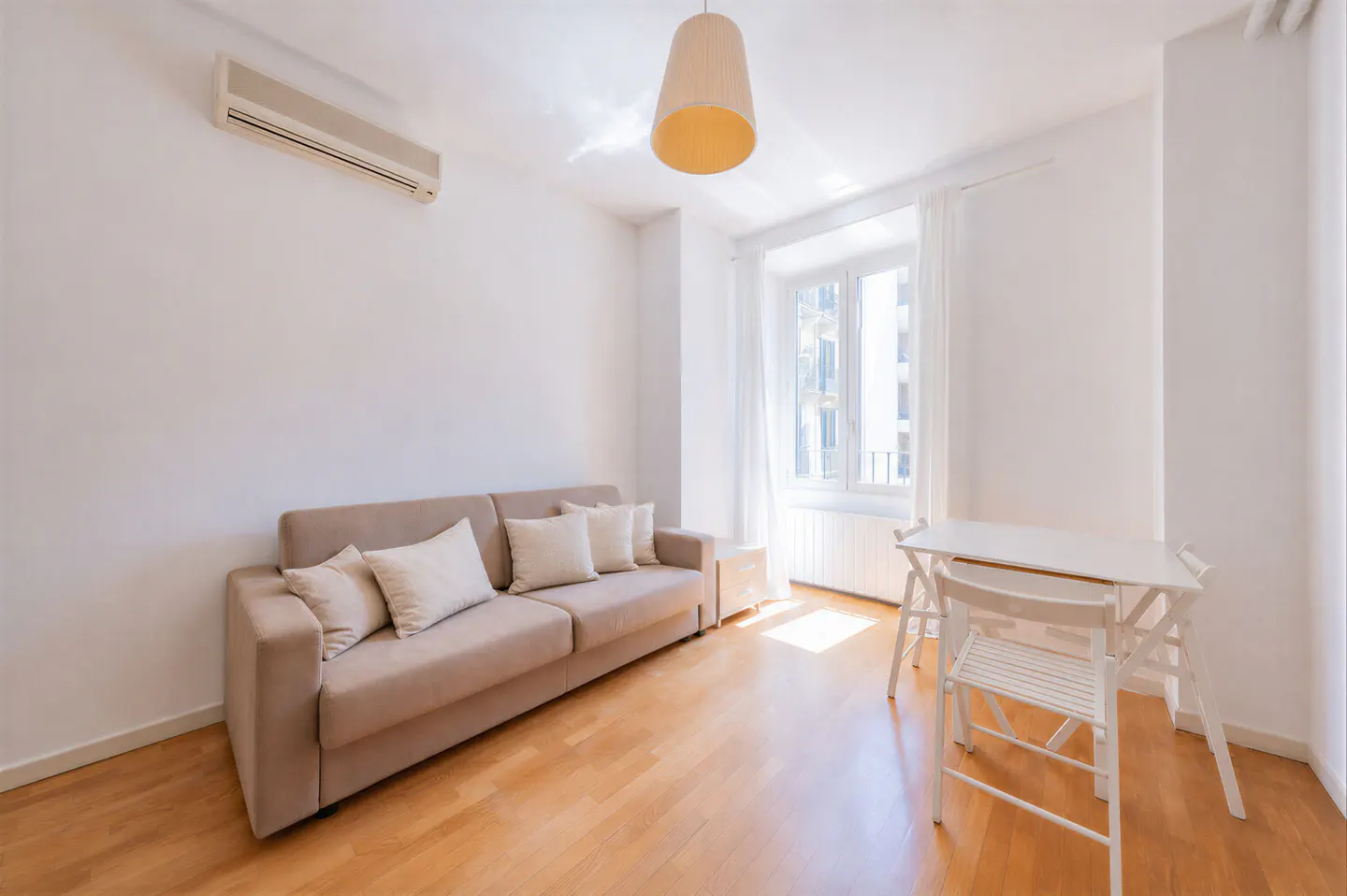 Bright living room with a beige sofa, white dining table and chairs, hardwood floors, and an air conditioning unit.