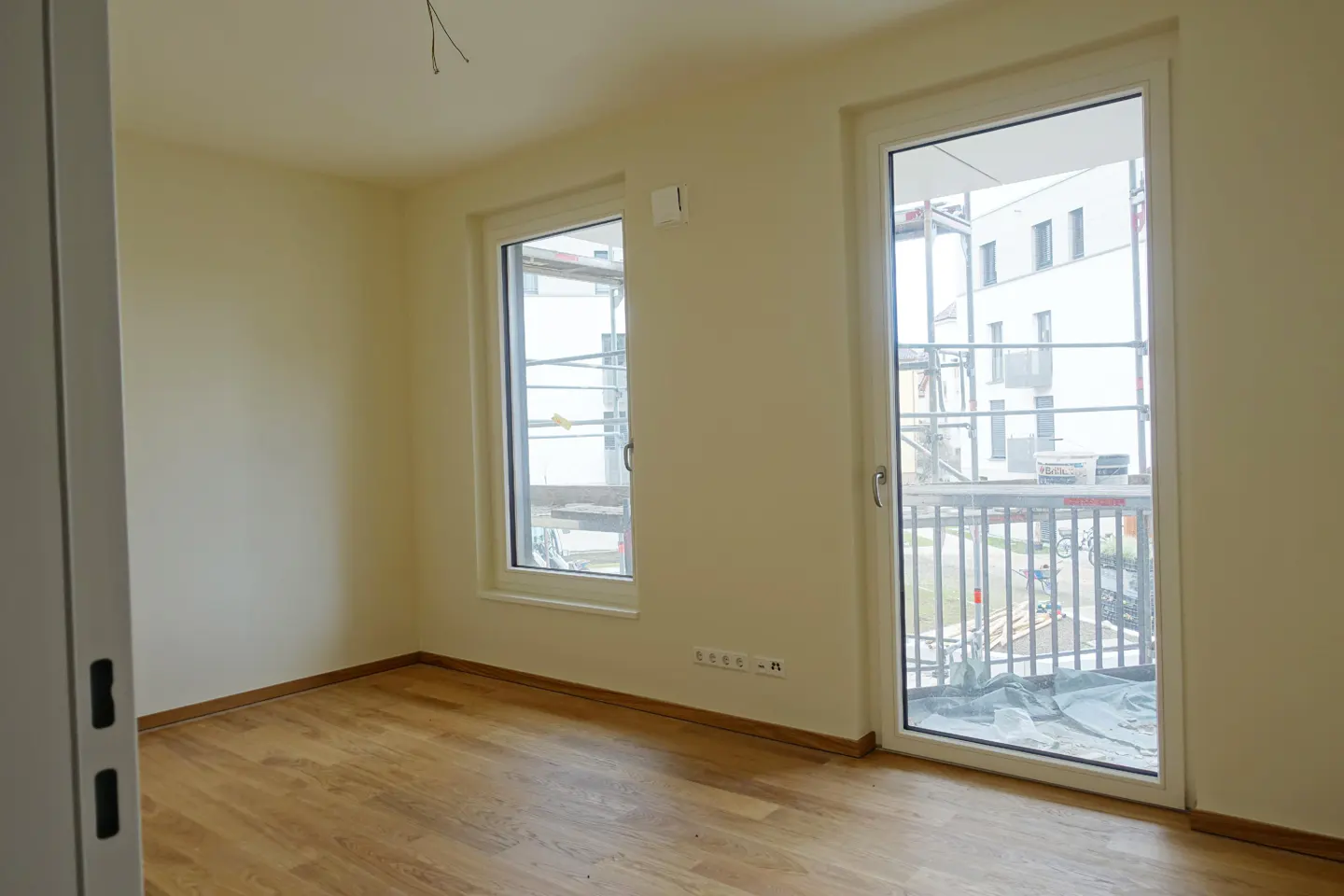 A bright, empty room with light wood floors, cream walls, and two white-framed windows. Outside, scaffolding is visible.