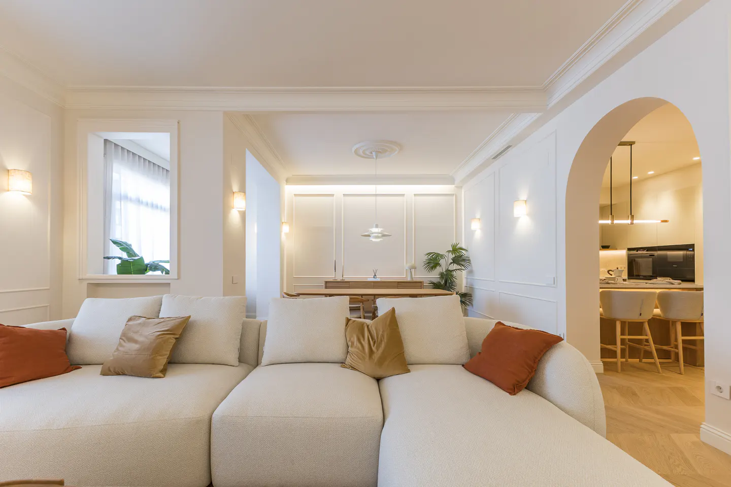 Bright, modern living room with a large cream sectional sofa, orange pillows, and an arched doorway leading to a kitchen.