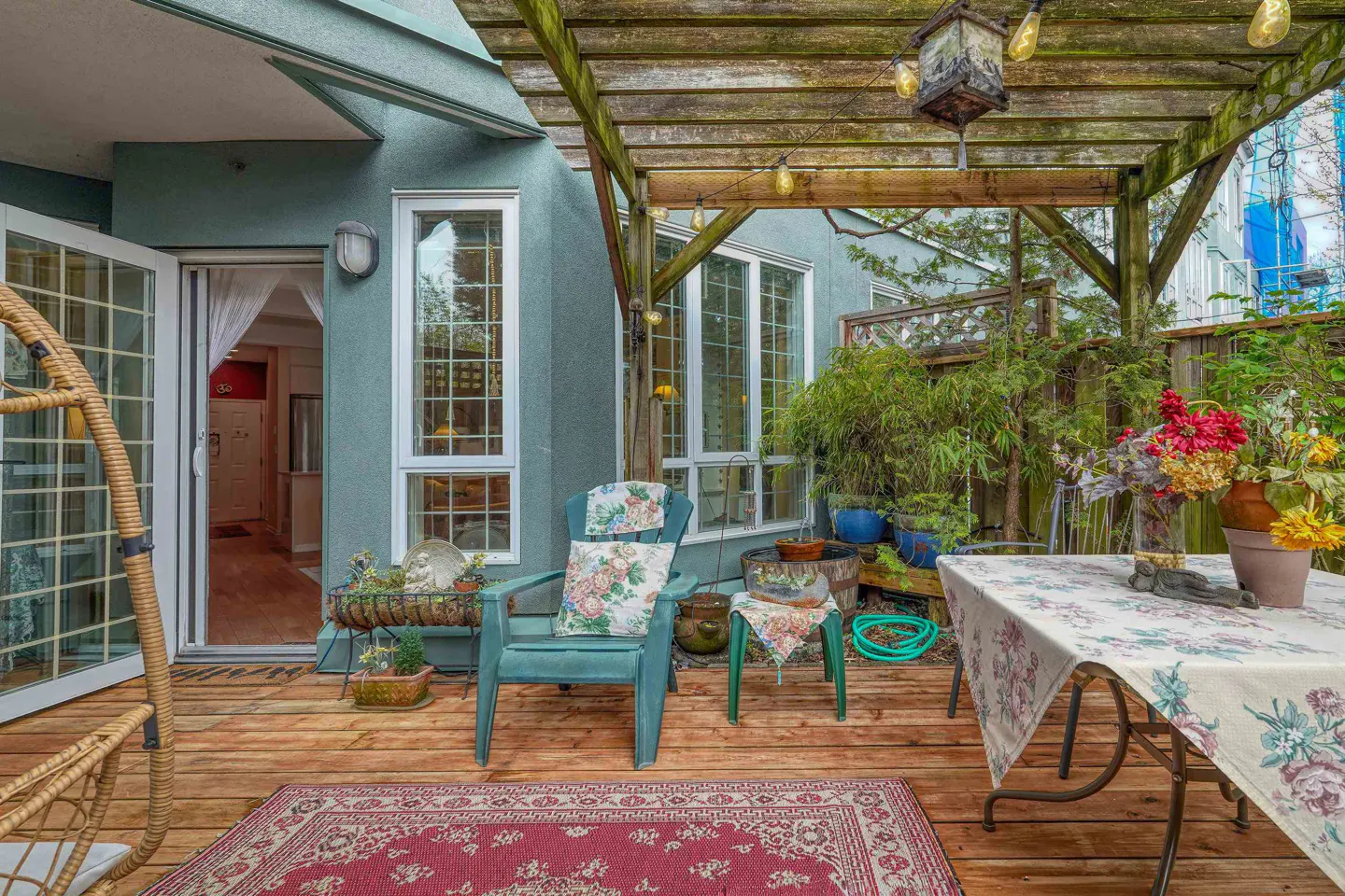 A wooden deck patio with a pergola, floral-cushioned chair, dining table, and a red patterned rug outside a home entrance.