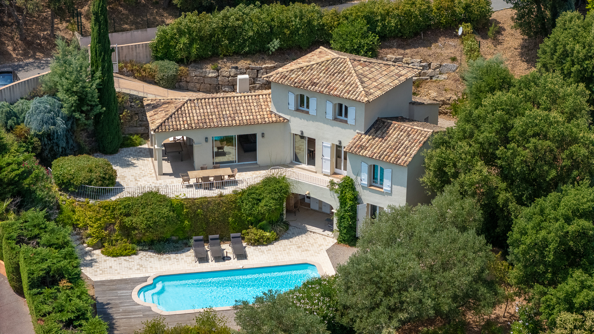Aerial view of a two-story beige villa with a tiled roof, outdoor dining patio, and a swimming pool surrounded by trees.