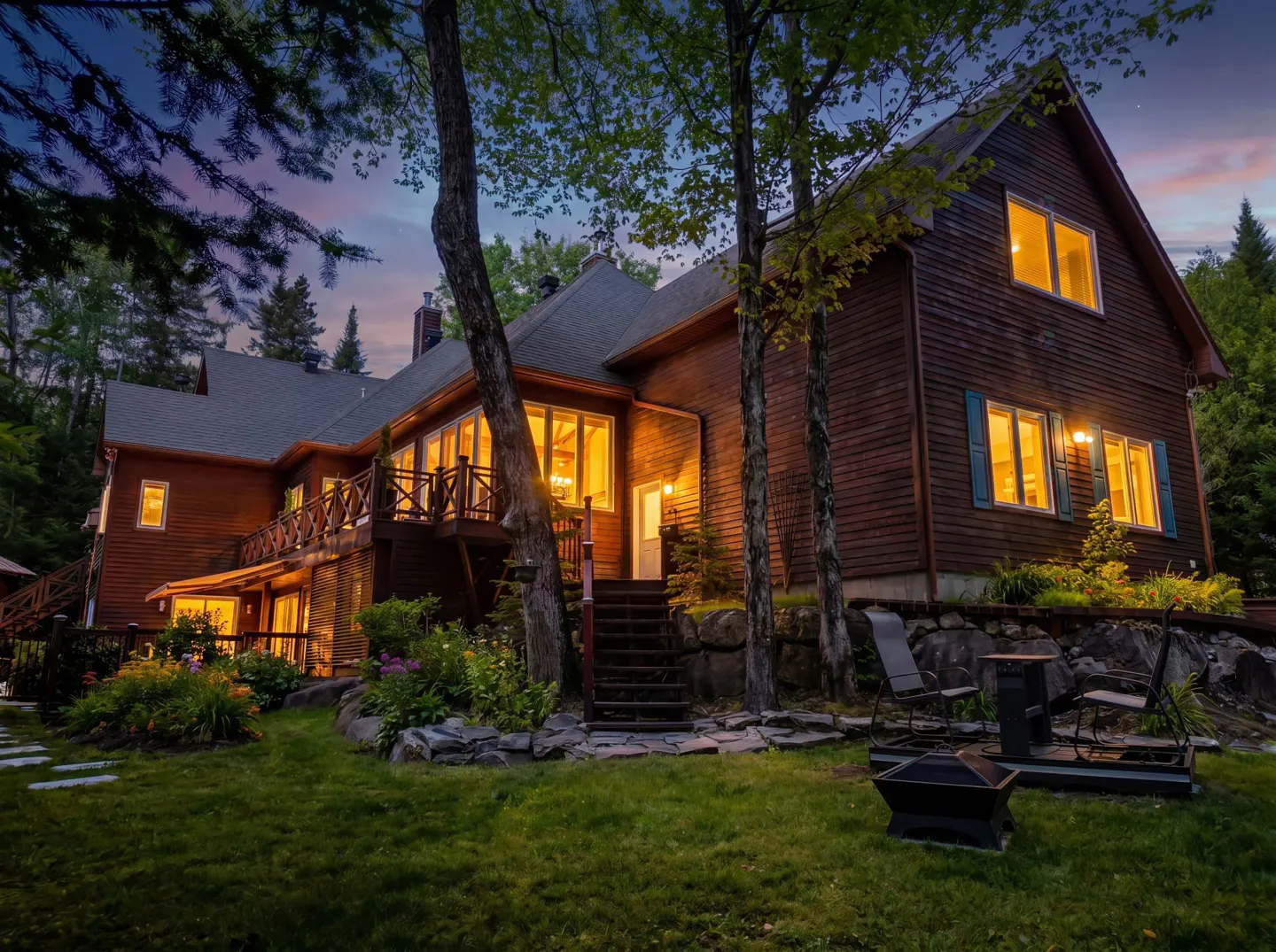A large, two-story brown house with many windows glowing with warm light at dusk. A green lawn and trees surround the house.