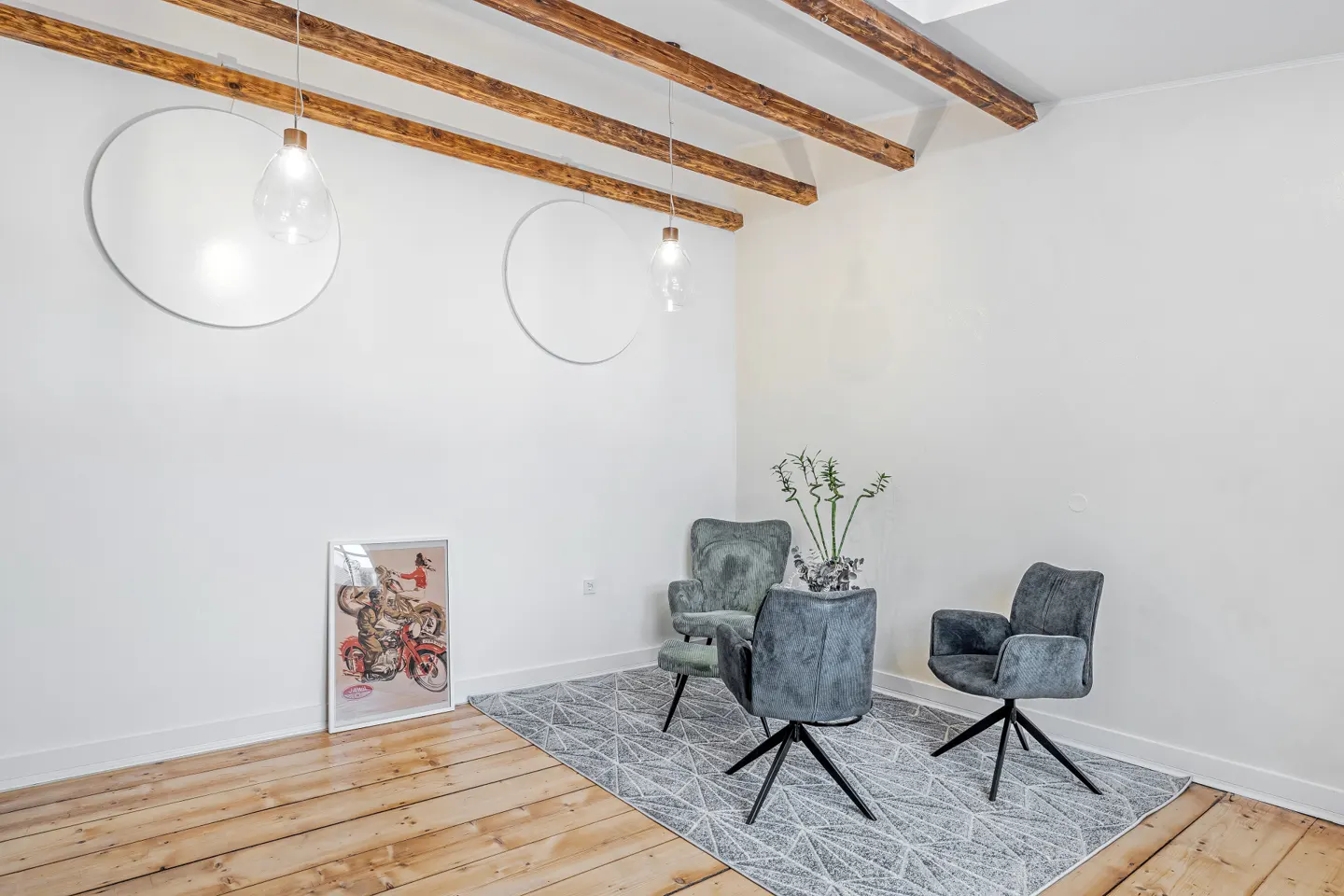 A bright room with wooden floors, white walls, and exposed beams. Three chairs sit on a geometric rug, with a framed print and round wall decor.