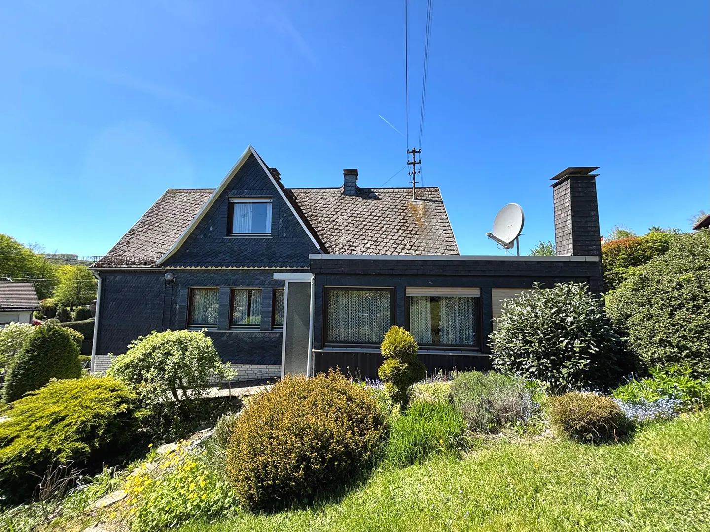 A gray house with a brown roof and a satellite dish sits on a green lawn under a blue sky.