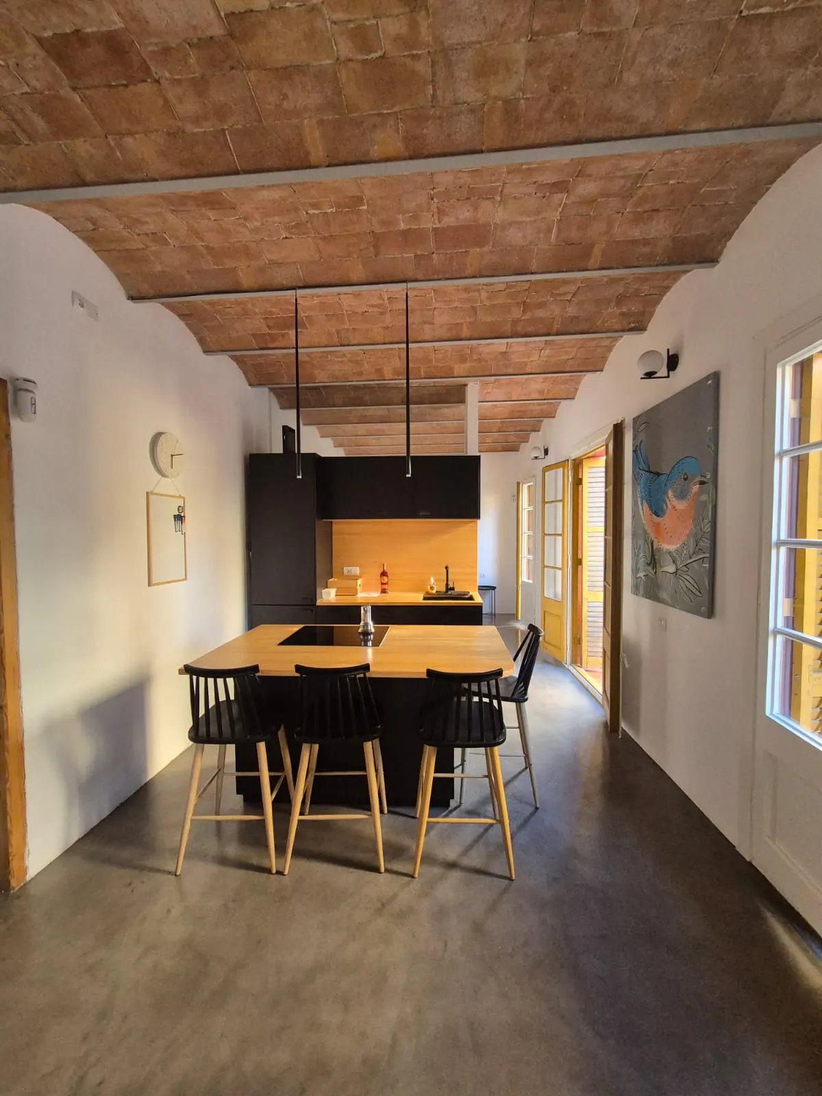 A modern kitchen with a brick ceiling, black cabinets, and a light wood island with black chairs. A painting of a bird hangs on the white wall.
