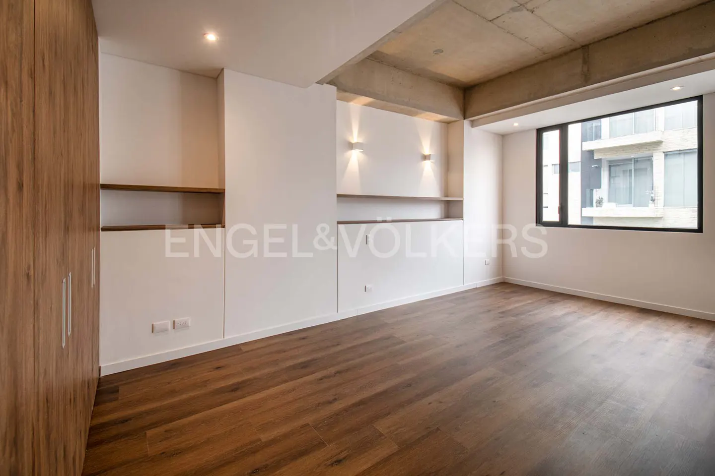 An empty bedroom with wood floors, a large window, and a wood closet. The walls are white with built-in shelves and sconces. The ceiling is concrete.