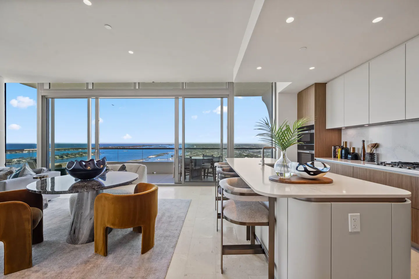 Open-concept condo interior with a kitchen island, dining table, and ocean view through sliding glass doors. Neutral tones and modern design.