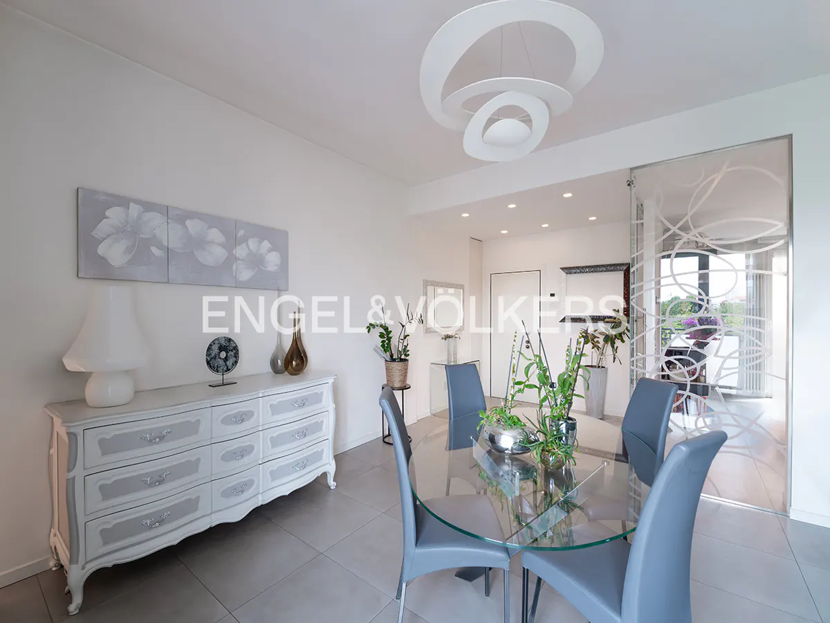 Bright dining room with a glass table, gray chairs, and a white dresser with a lamp. A modern white chandelier hangs above.