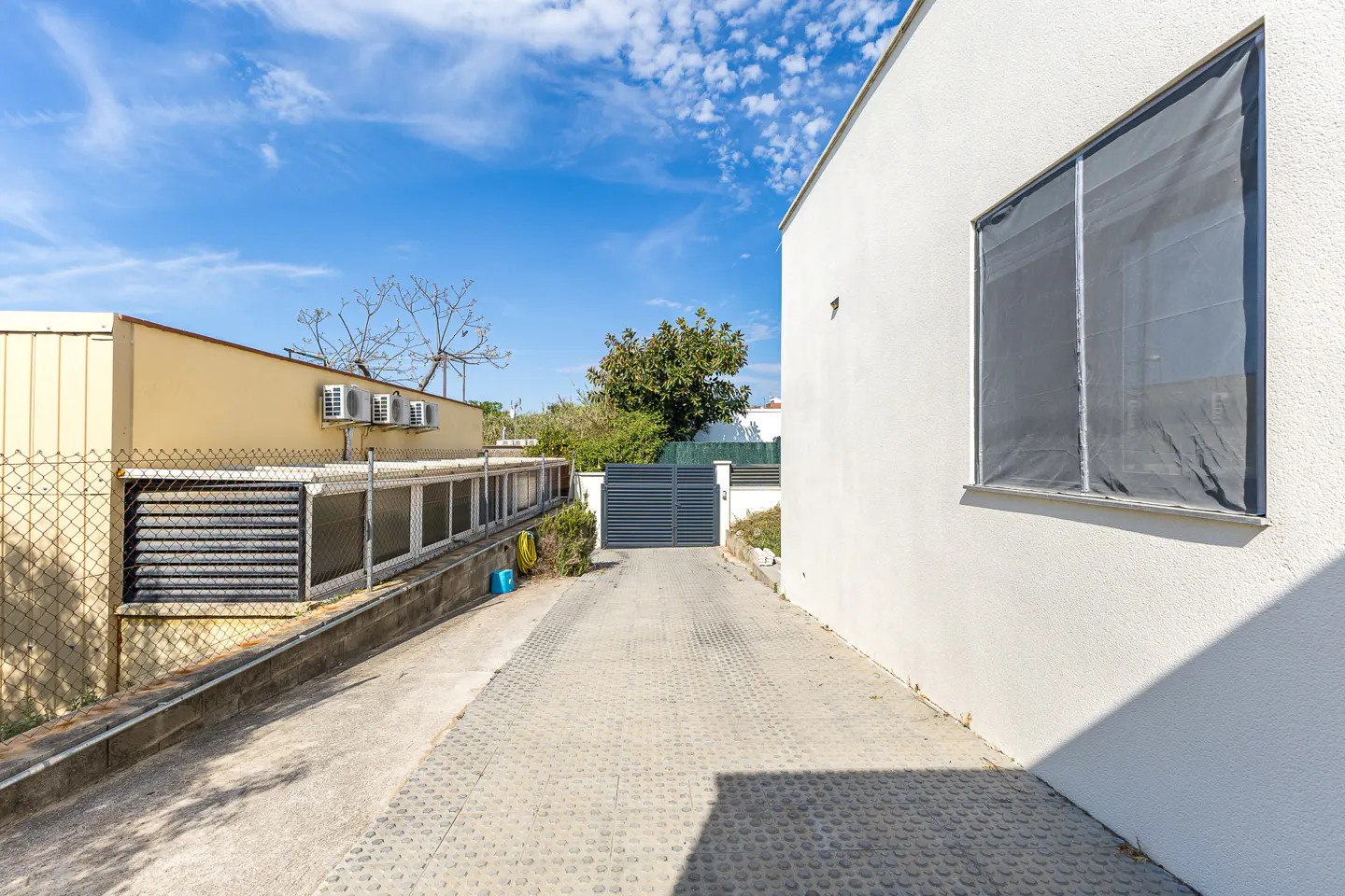 Exterior view of a property with a gray driveway leading to a gate, a white building on the right, and a yellow building on the left.