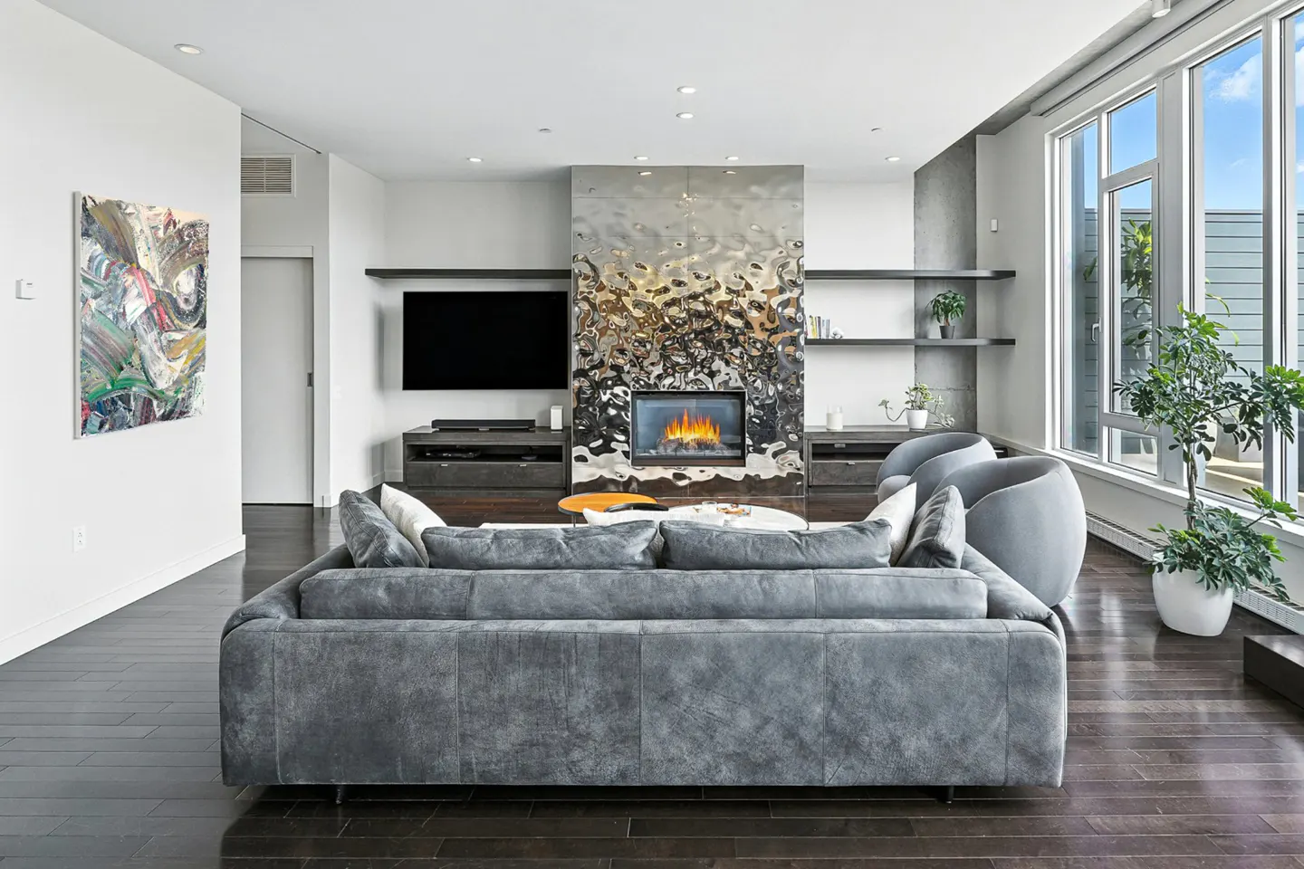Living room with gray sofa, fireplace with textured metal surround, TV, and large windows. Dark wood floors.