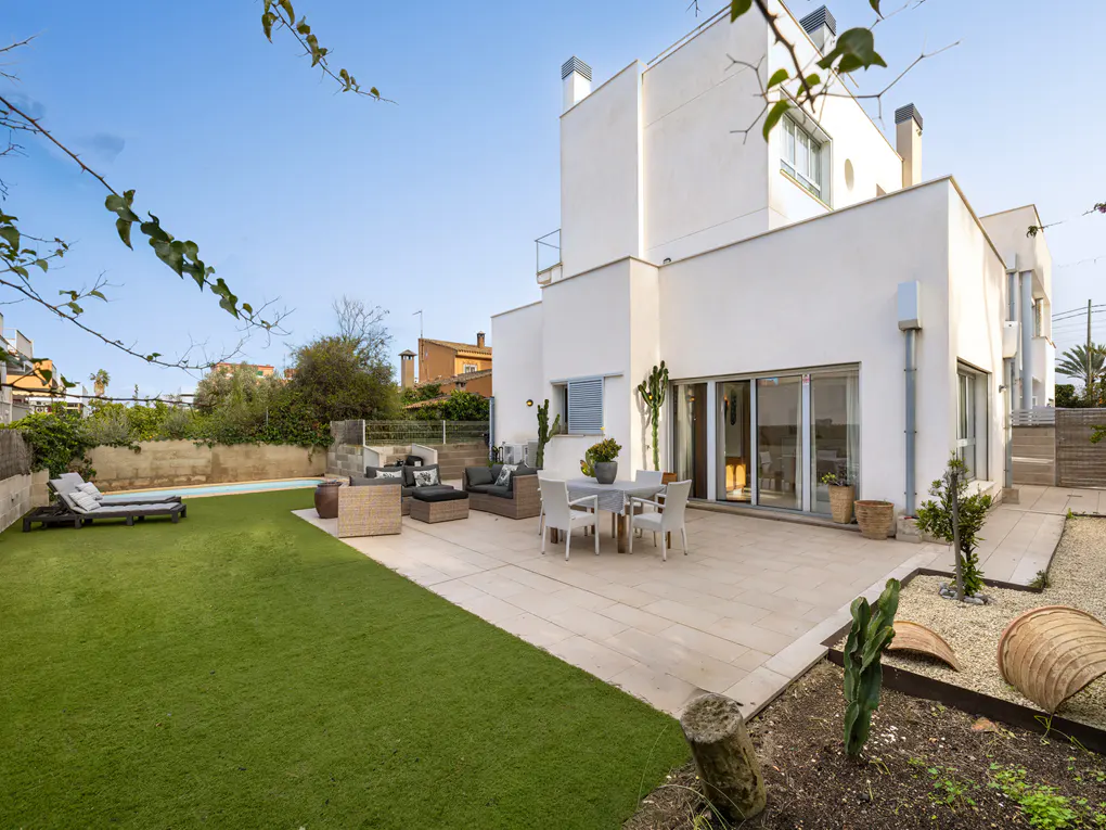 Backyard of a modern white house with a patio, lawn, pool, and outdoor furniture.