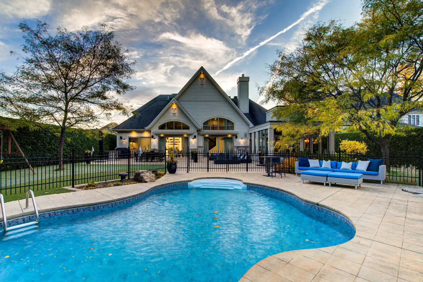 Backyard view of a two-story house with a pool, patio, and black metal fence. Blue skies with clouds overhead.