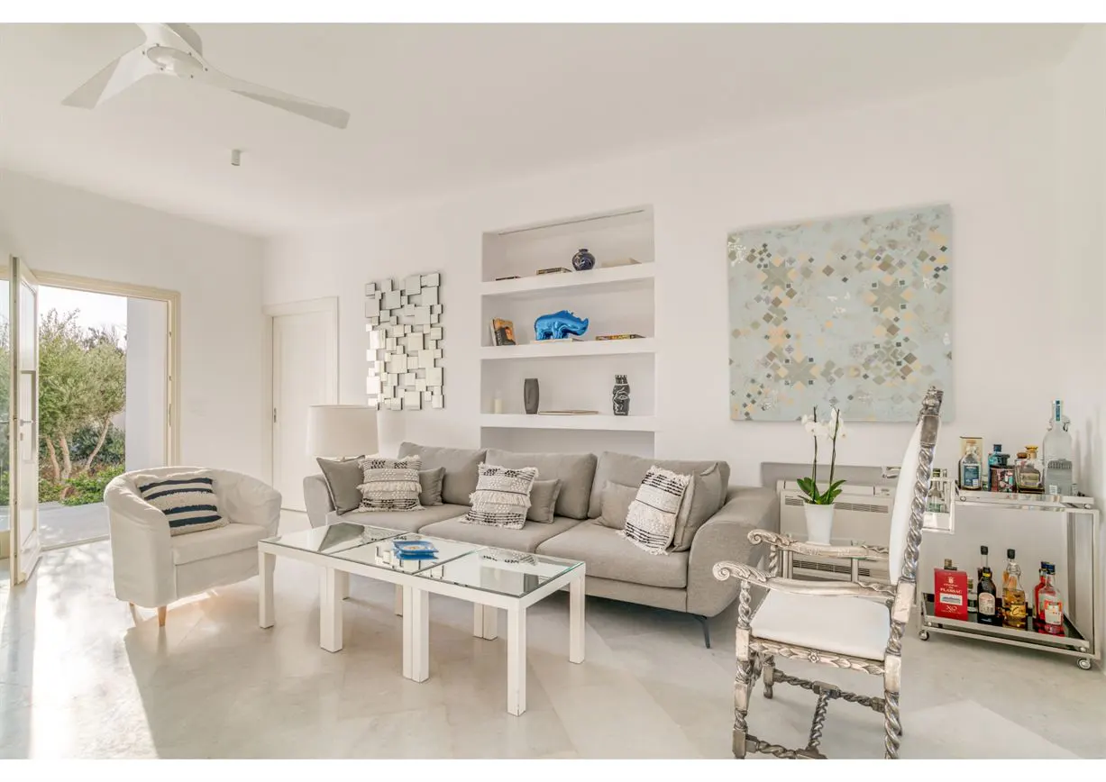 Bright living room with white walls, a gray sofa, and a glass coffee table. Shelves display decor, and a bar cart holds bottles. An open door leads to greenery.