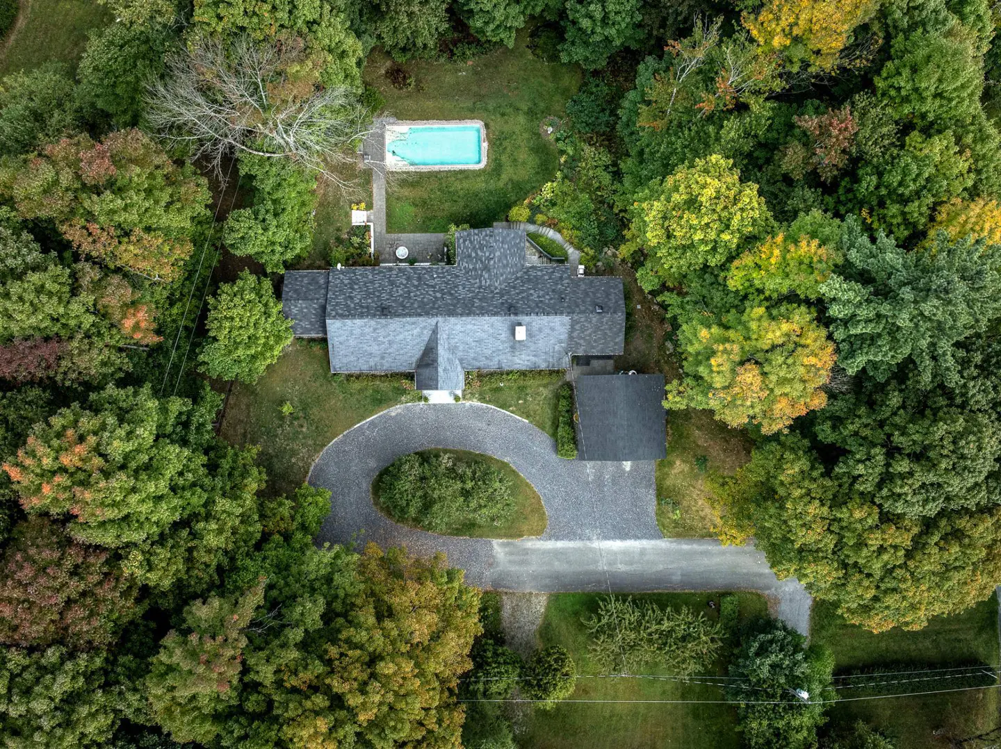 Aerial view of a house with a gray roof, surrounded by green trees, a circular driveway, and a blue swimming pool.