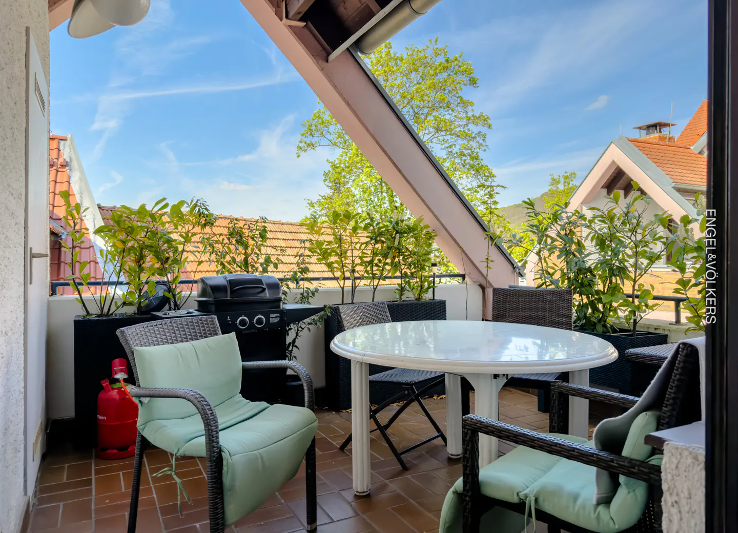 Balcony with a white table, chairs with light green cushions, a grill, and plants in black planters. Rooftops and blue sky in the background.