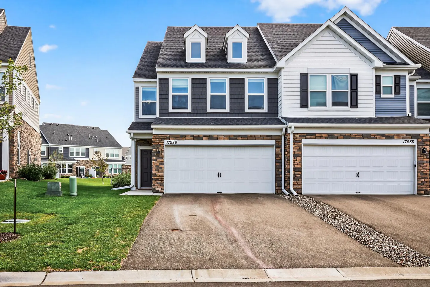 Two-story townhouses with white garage doors, stone and gray siding, and dormer windows under a blue sky.