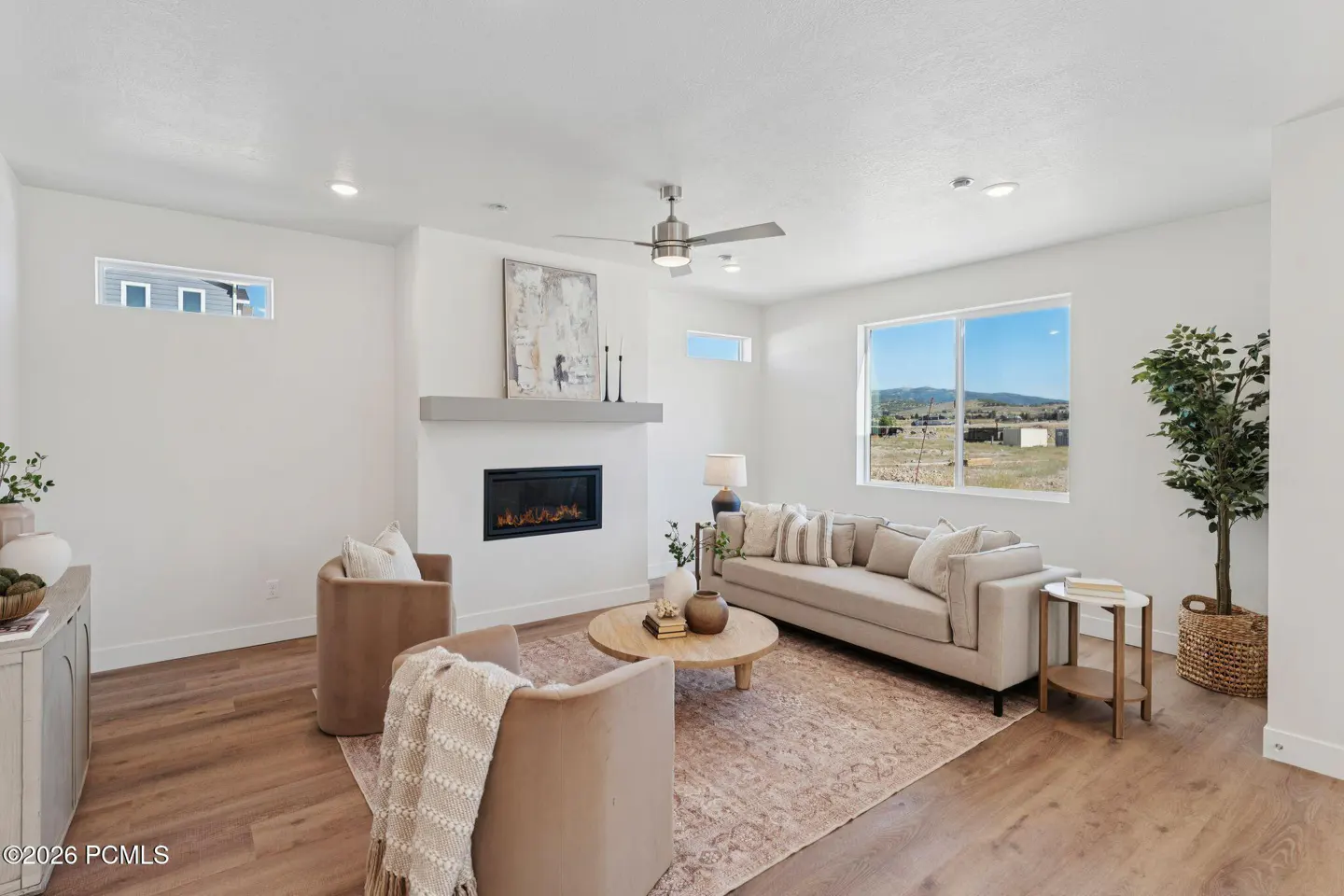 Modern living room with a fireplace, beige sofa, two armchairs, a round coffee table, and a view of hills through a window.