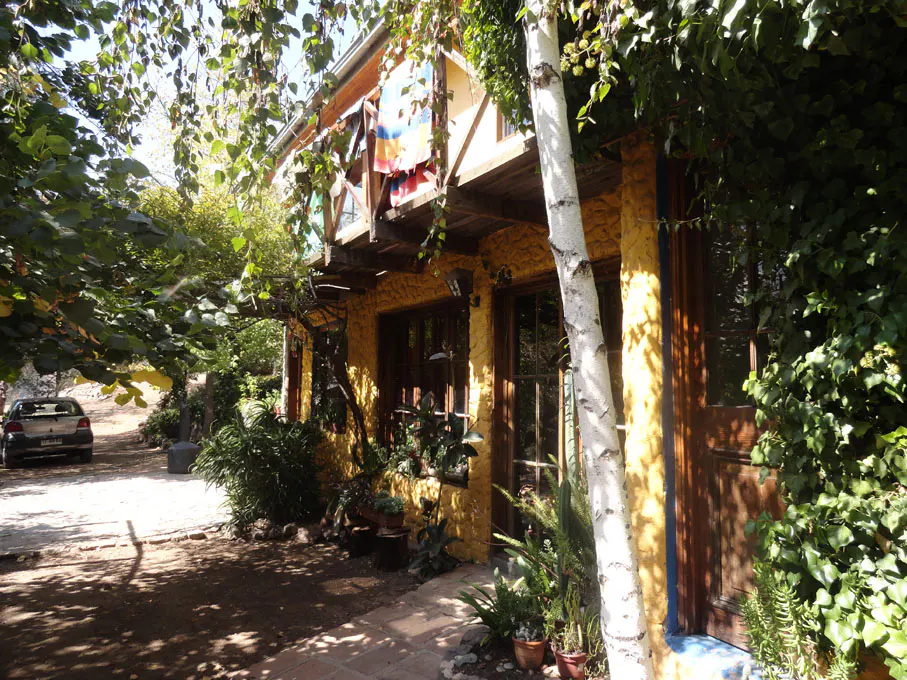Exterior view of a yellow stone house with brown trim, surrounded by lush greenery and a birch tree. A car is parked in the driveway.