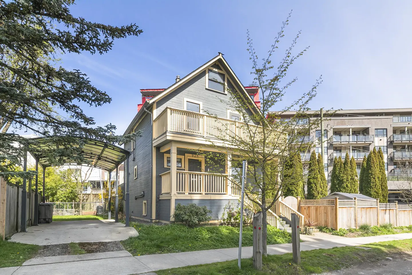 Two-story gray house with a tan porch and balcony, a carport, and a green lawn under a blue sky.