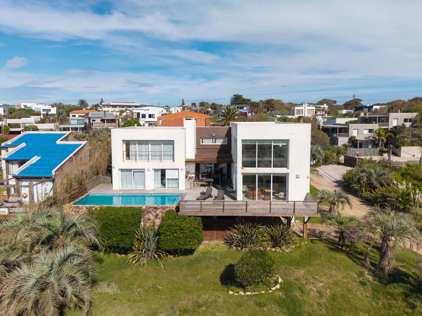 Two-story modern white house with a pool and wooden deck, surrounded by green lawn and palm trees under a blue sky.