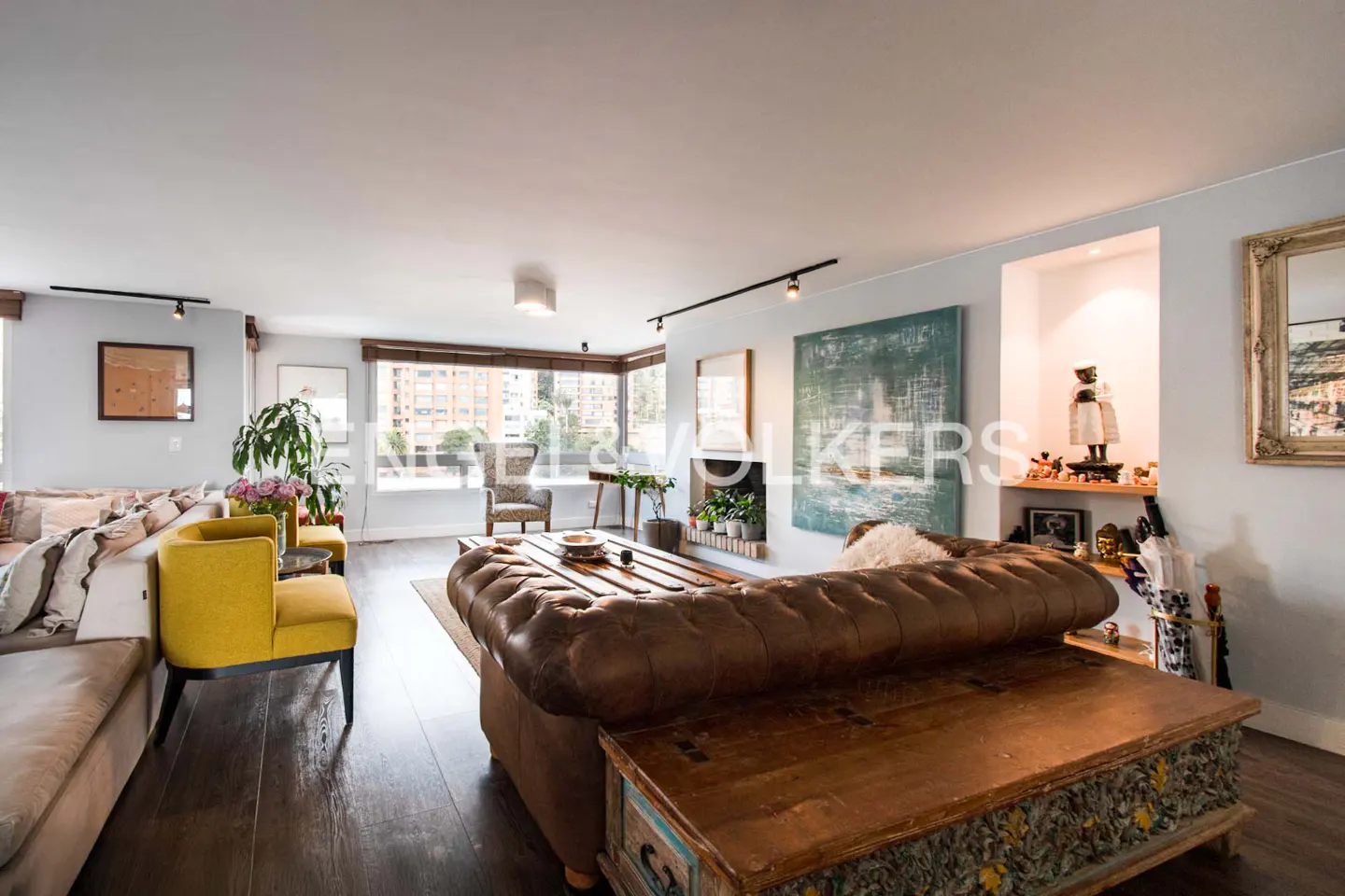 A living room with a brown leather sofa, yellow chairs, and dark wood floors. A large window overlooks a city.