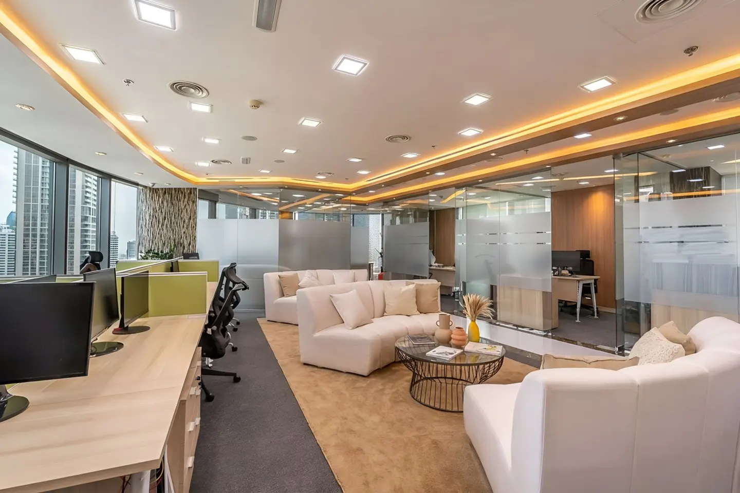 Bright office space with beige sofas, a round coffee table, and desks with computers. Large windows show a city view.