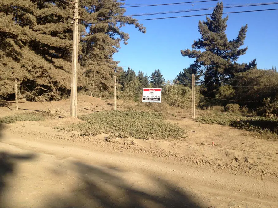 Land for sale with a "Vende" sign by Engel & Volkers, surrounded by trees and a wire fence on a sunny day.