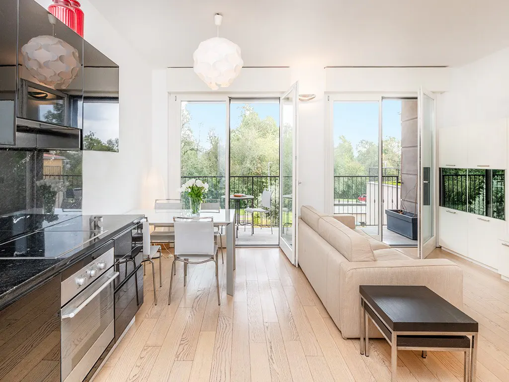 Bright, modern apartment interior with a kitchen, dining table, and living area leading to a balcony with green trees.