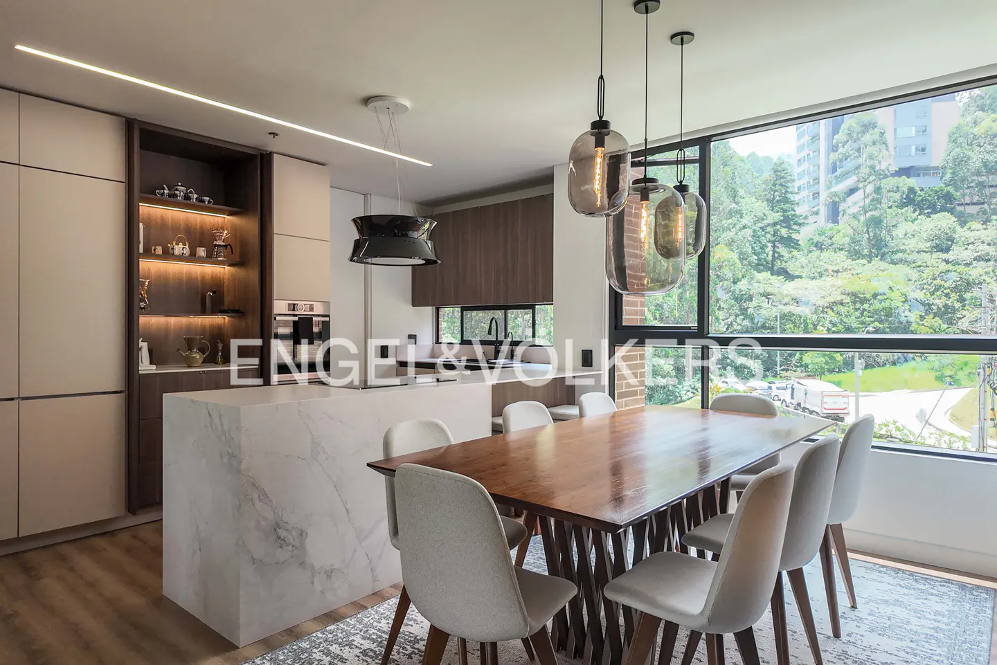 A modern dining area with a wooden table, white chairs, and pendant lights. Large windows offer a view of lush greenery.