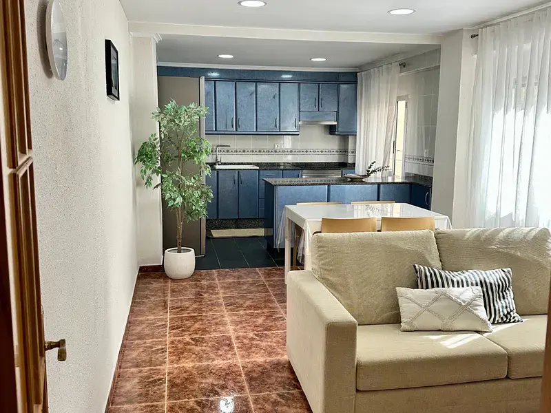 A bright, open-concept living space with a beige sofa, brown tile floor, and blue kitchen cabinets. A potted plant sits near the kitchen entrance.