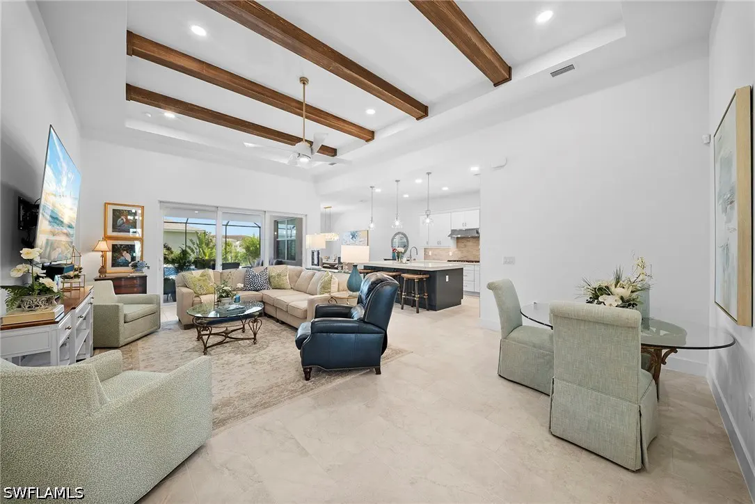 Open-concept living room with wood-beamed tray ceiling, beige sectional, dark blue recliner, and dining area with glass table.