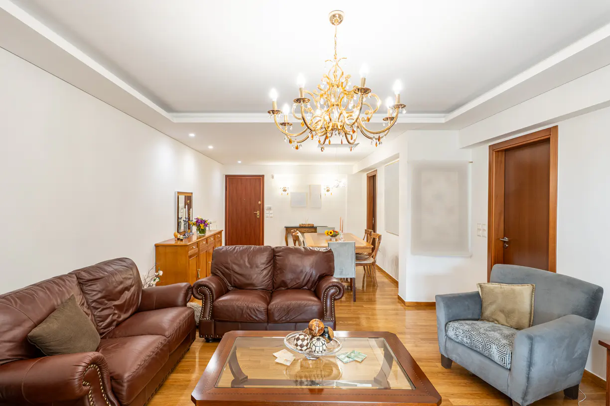 Bright living room with brown leather sofas, a gray armchair, and a wooden coffee table. A gold chandelier hangs from the white ceiling.