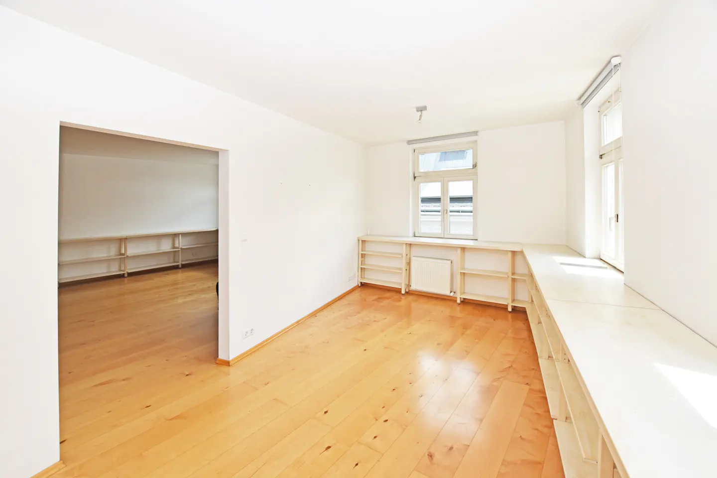 Bright, empty room with light wood floors and white walls. Built-in shelves line the walls, and a window lets in natural light.