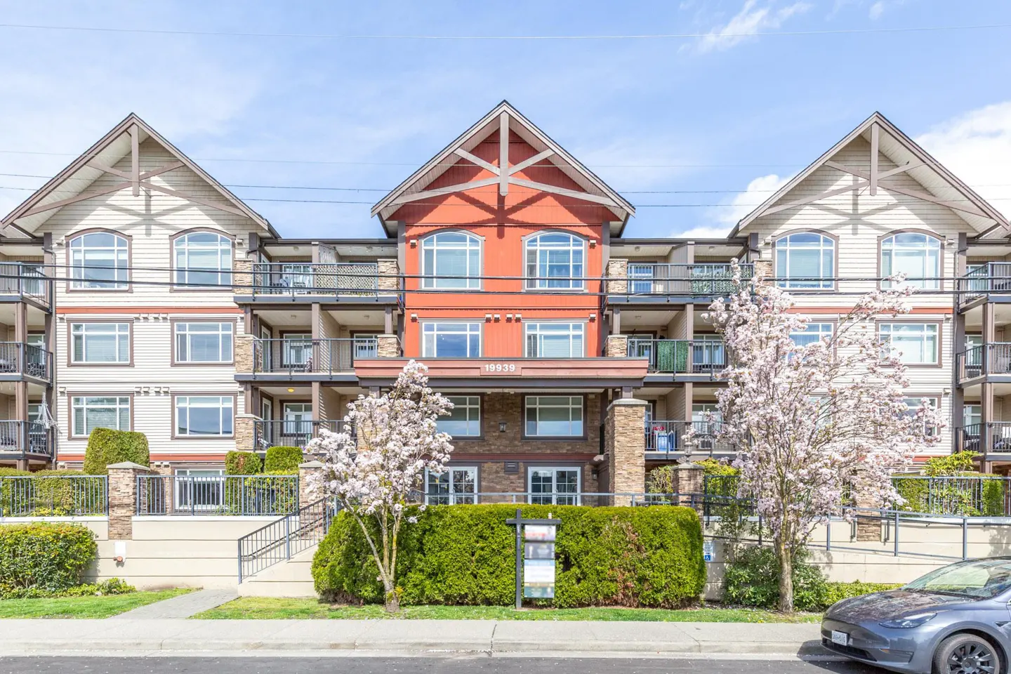 Exterior of a three-story apartment building with balconies, red and beige siding, and blooming trees in front.