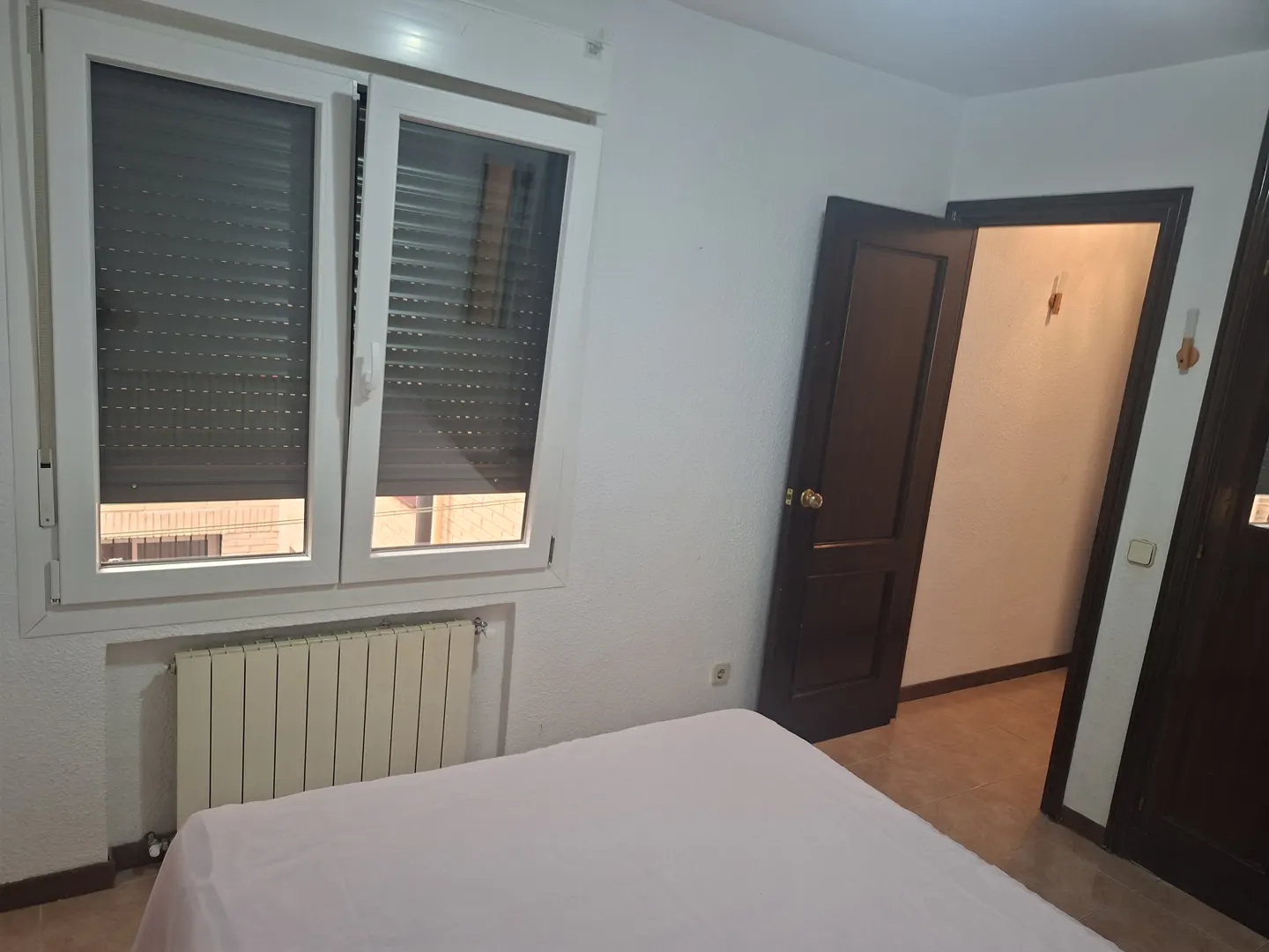Bedroom with white walls, a bed with a pink sheet, a window with closed blinds, and an open dark brown door.