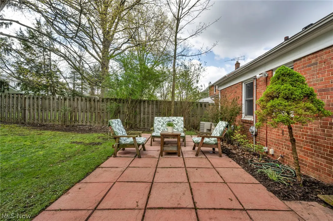 Backyard patio with red pavers, wooden furniture with leaf-patterned cushions, and a green lawn with a wooden fence.