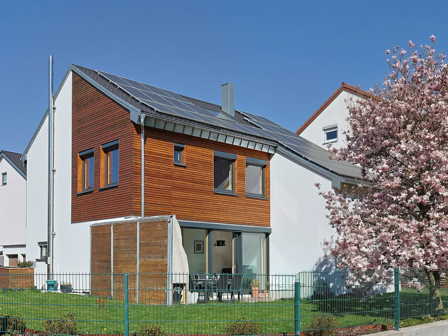 Two-story house with wood and white siding, solar panels, and a green lawn with a metal fence. A flowering tree is on the right.