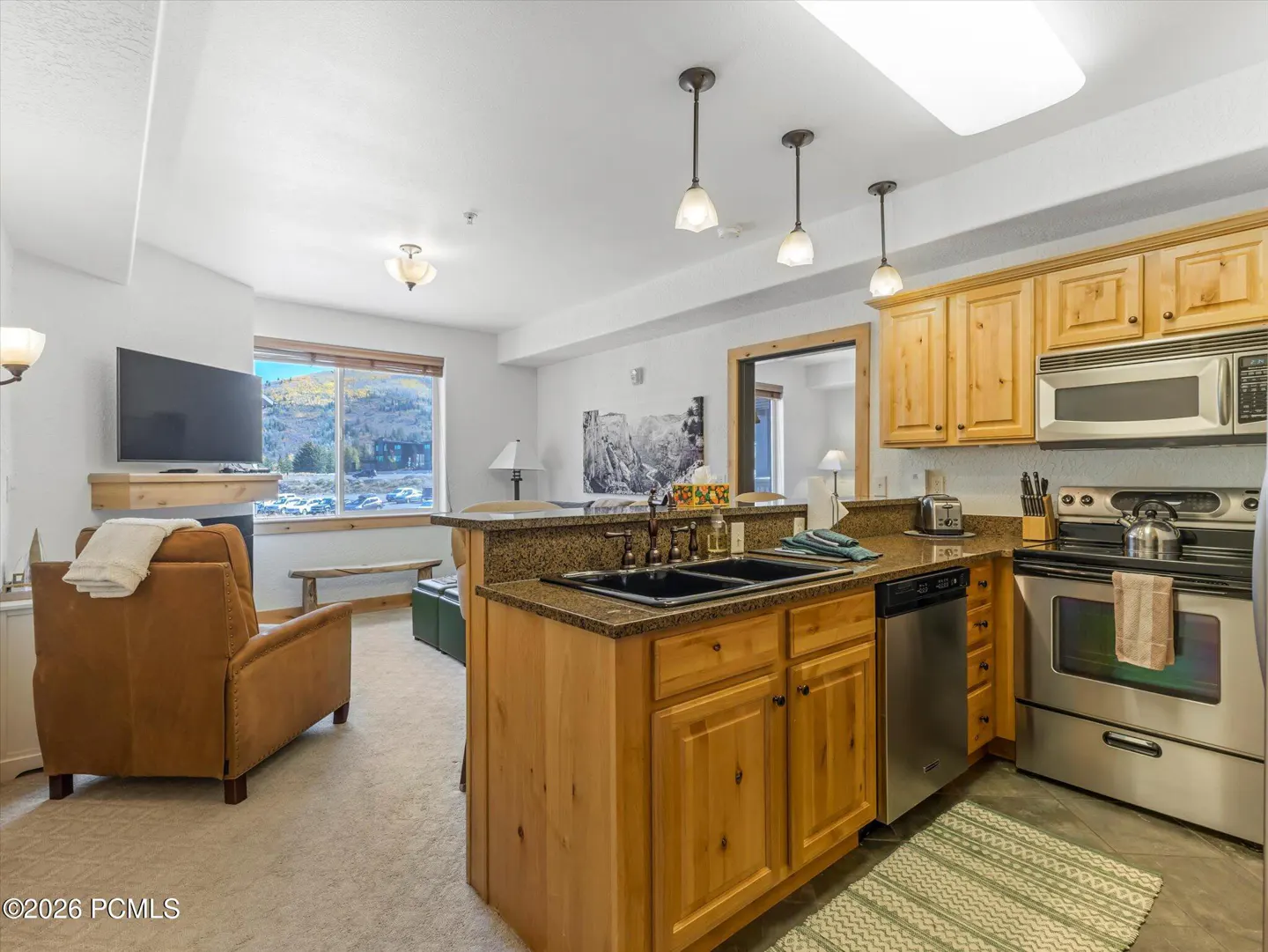 Condo interior with a kitchen island, stainless steel appliances, and a living room with a TV and a brown leather armchair.