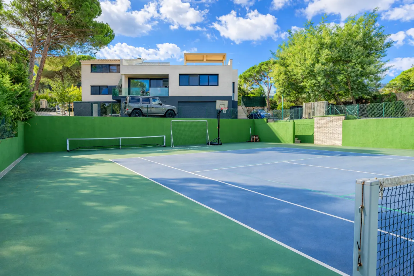 A modern house overlooks a blue and green tennis court with a basketball hoop and soccer net.