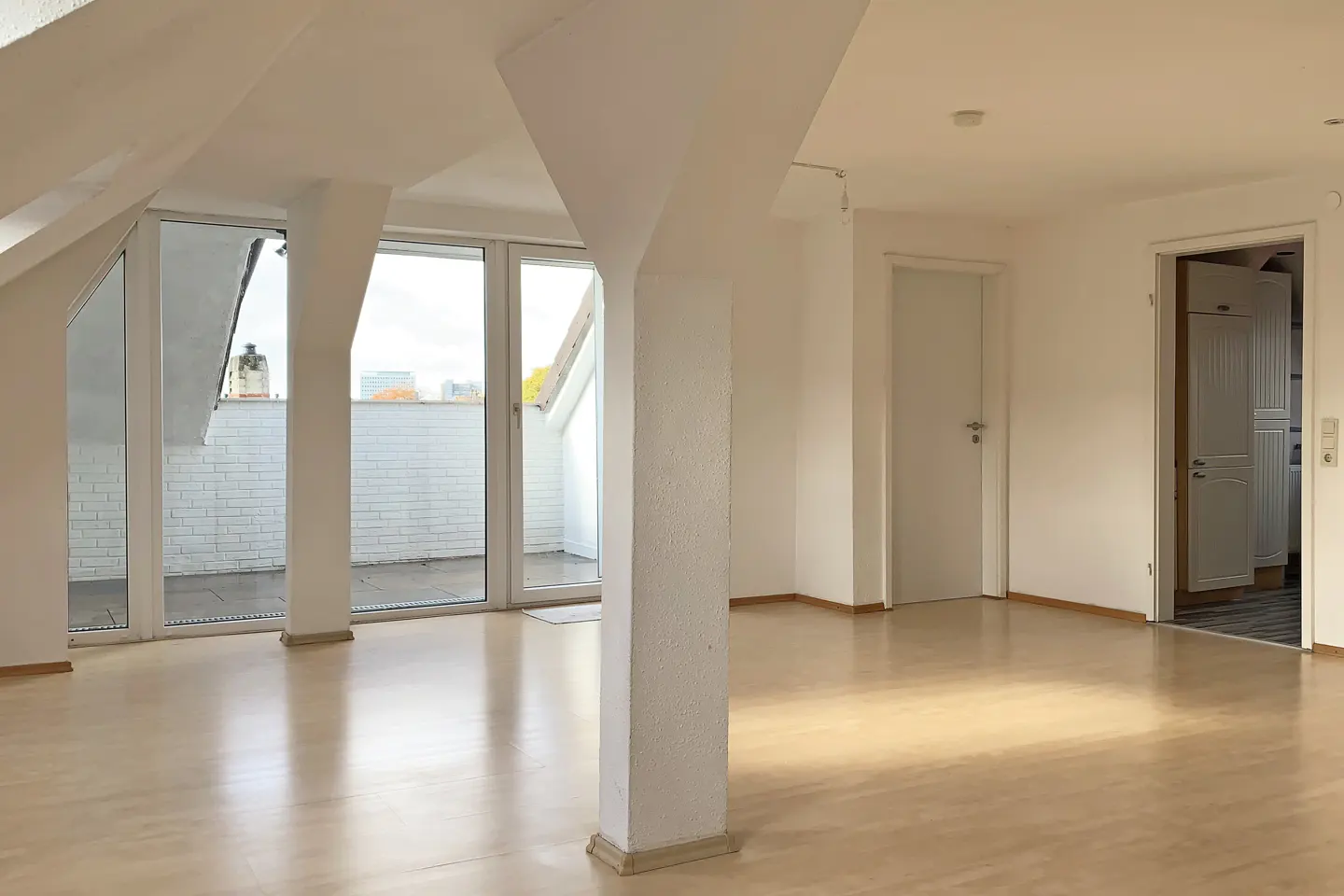 Bright, empty room with light wood floors, white walls, and a large white support column. Glass doors lead to a balcony.