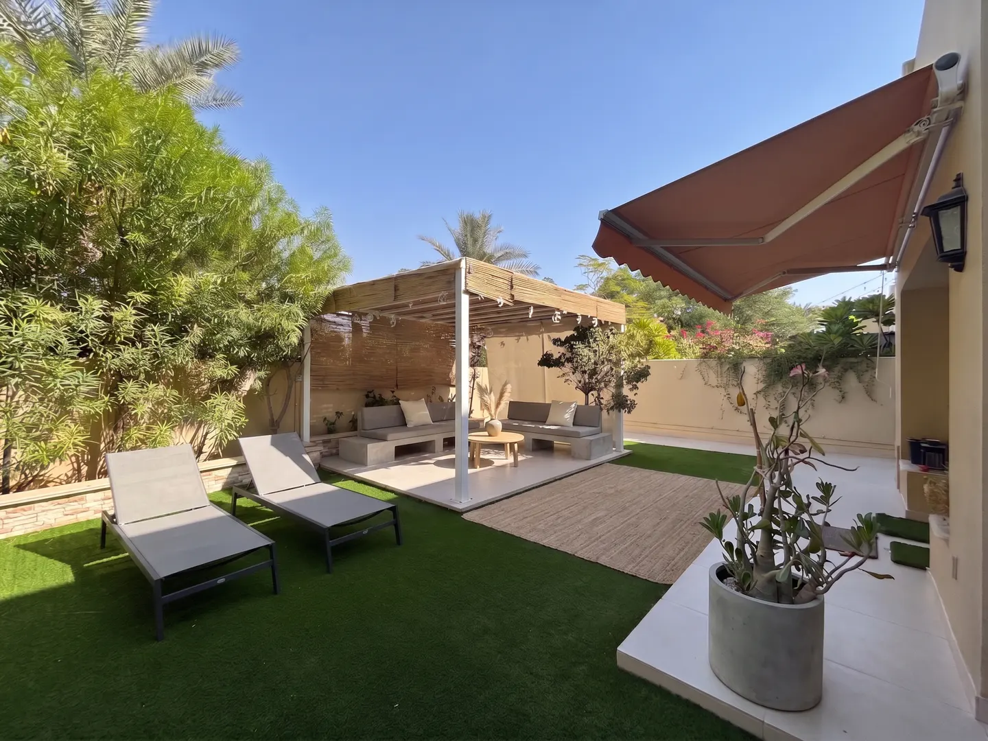Sunny backyard with artificial grass, two lounge chairs, a shaded pergola seating area, and a potted plant on the patio.