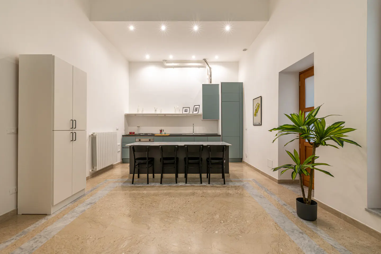 A modern kitchen with white walls, light-colored marble floors, and sage green cabinets. A black island with four chairs sits in the center. A potted plant is near a window.