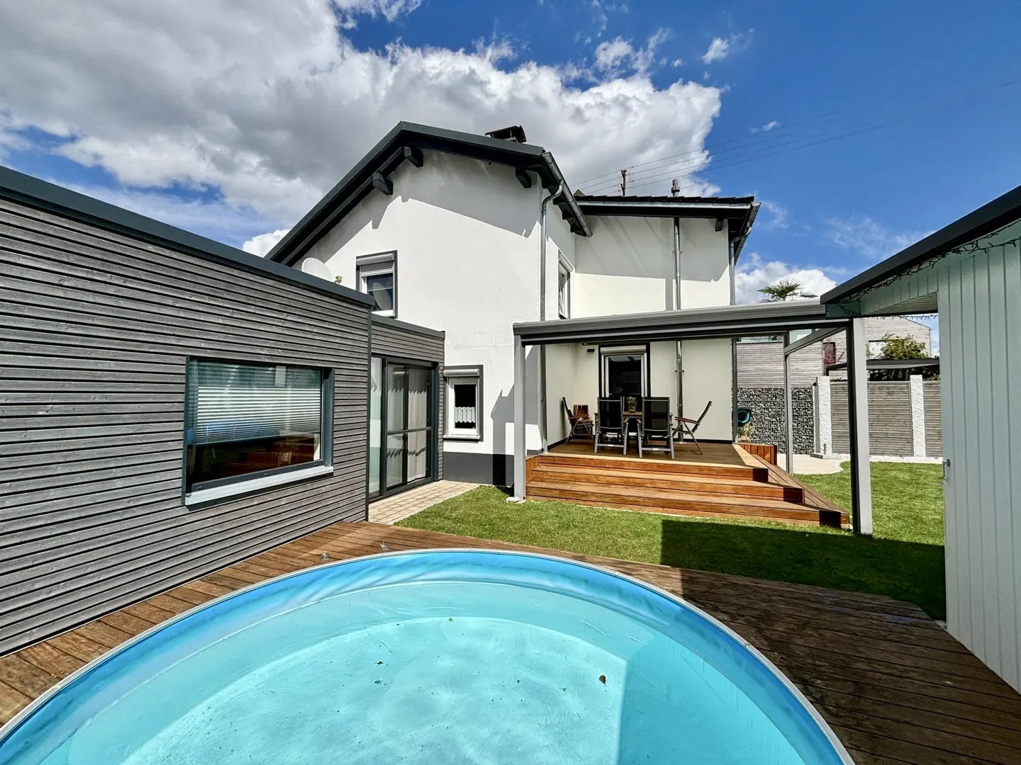 Backyard view of a white house with a wooden deck, a blue pool, and a gray wood-paneled building under a partly cloudy sky.