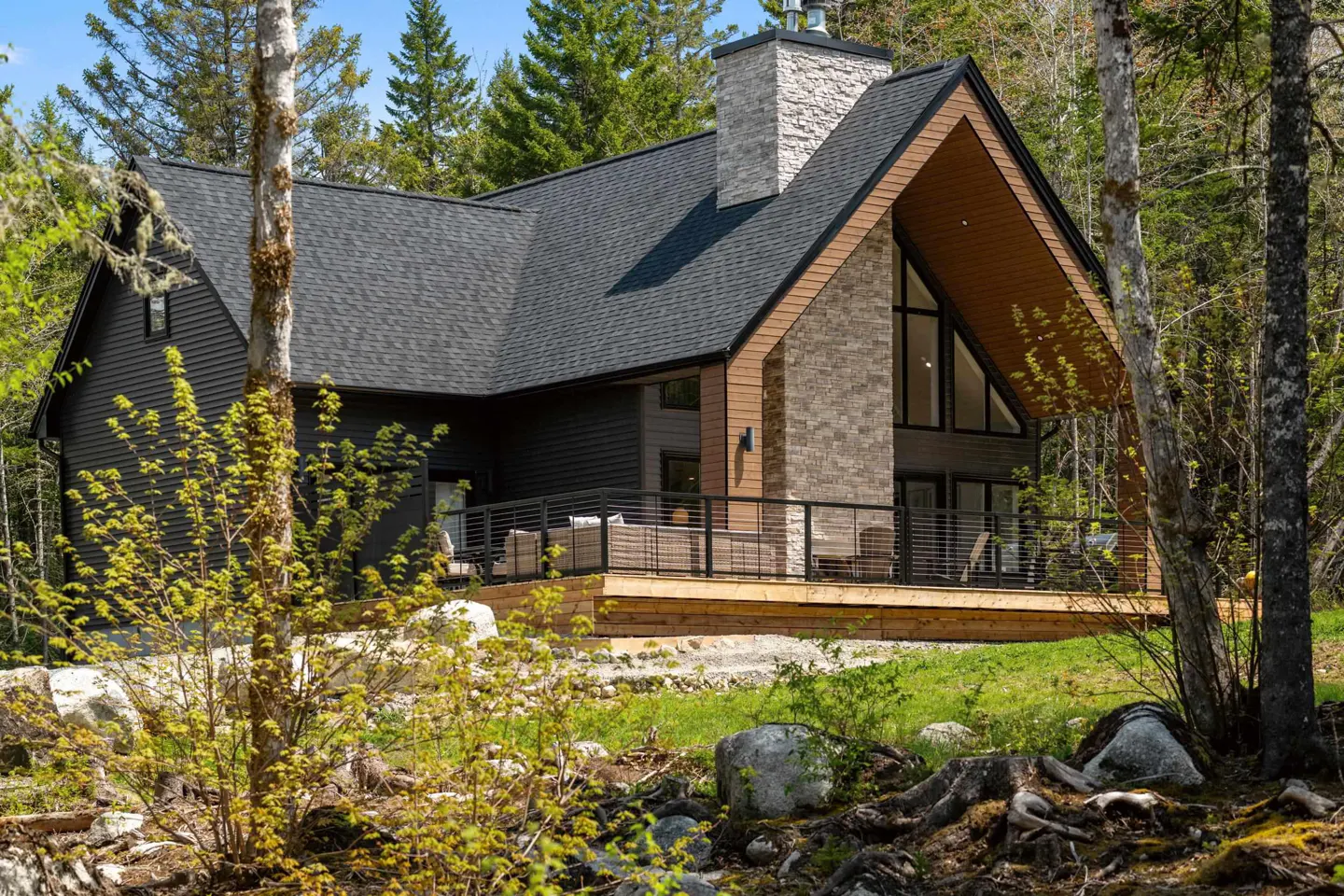 Modern black A-frame house with a stone chimney, wooden deck, and patio furniture, surrounded by a lush forest.