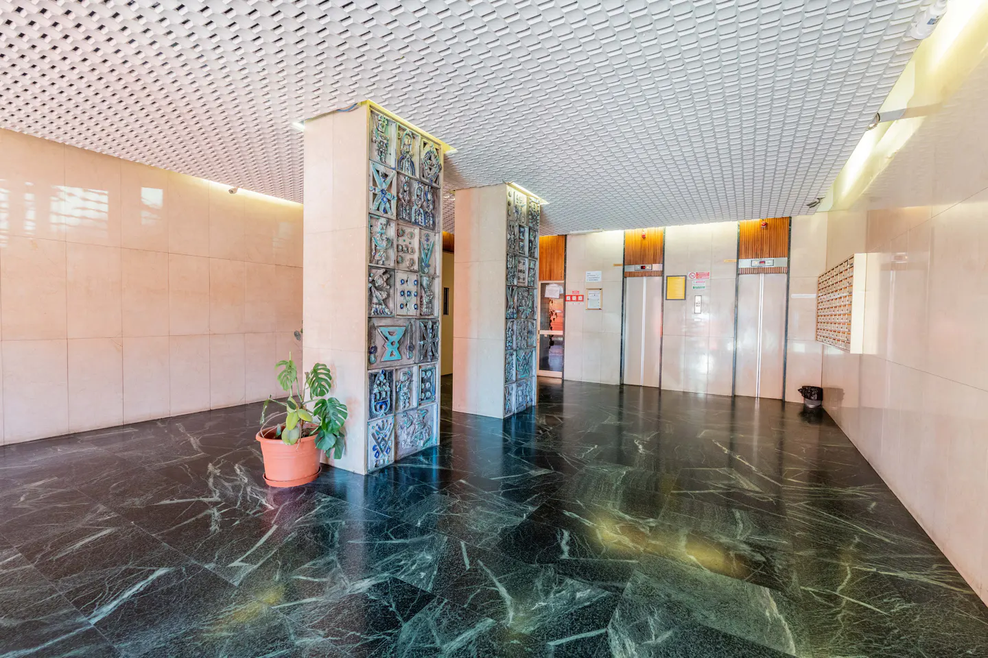 Lobby with green marble floor, tiled walls, and a white grid ceiling. Columns with decorative tiles and a potted plant add detail. Elevators are visible in the background.