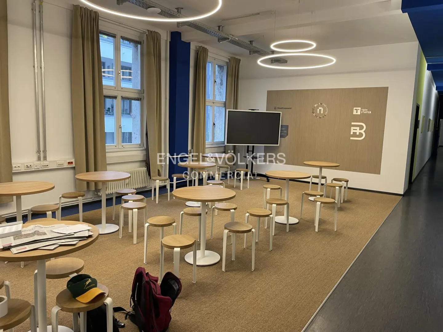 Bright room with round tables and stools, a large screen, and logos on the wall. Windows with beige curtains and circular lights overhead.