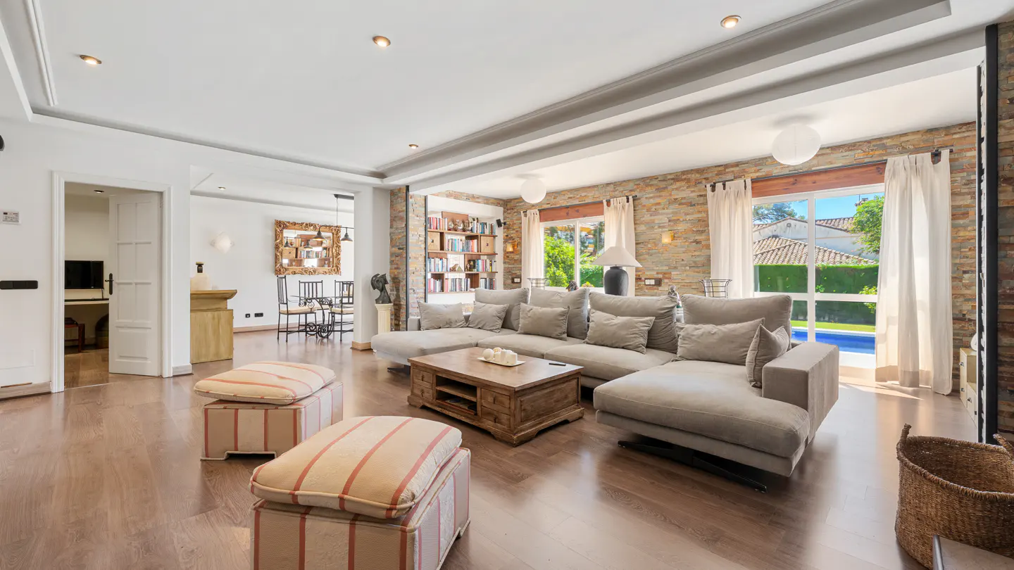Bright living room with a large gray sectional sofa, wooden coffee table, and striped ottomans on a wood floor. A stone wall and pool view are visible.