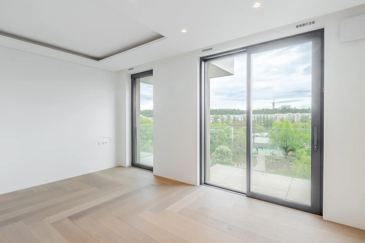 Bright, empty room with light wood floors, white walls, and large sliding glass doors with a city view.